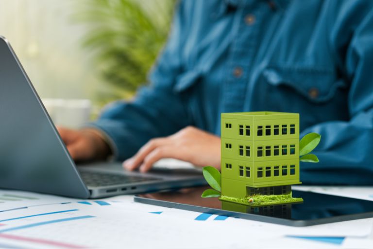 Hombre trabajando en su ordenador corporativo junto a una maqueta de un edificio verde, simbolizando sostenibilidad y comunicación empresarial.