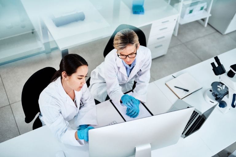 Dos investigadoras con batas de laboratorio revisan documentos y datos en una pantalla de computadora, asegurando la precisión de las traducciones médicas.